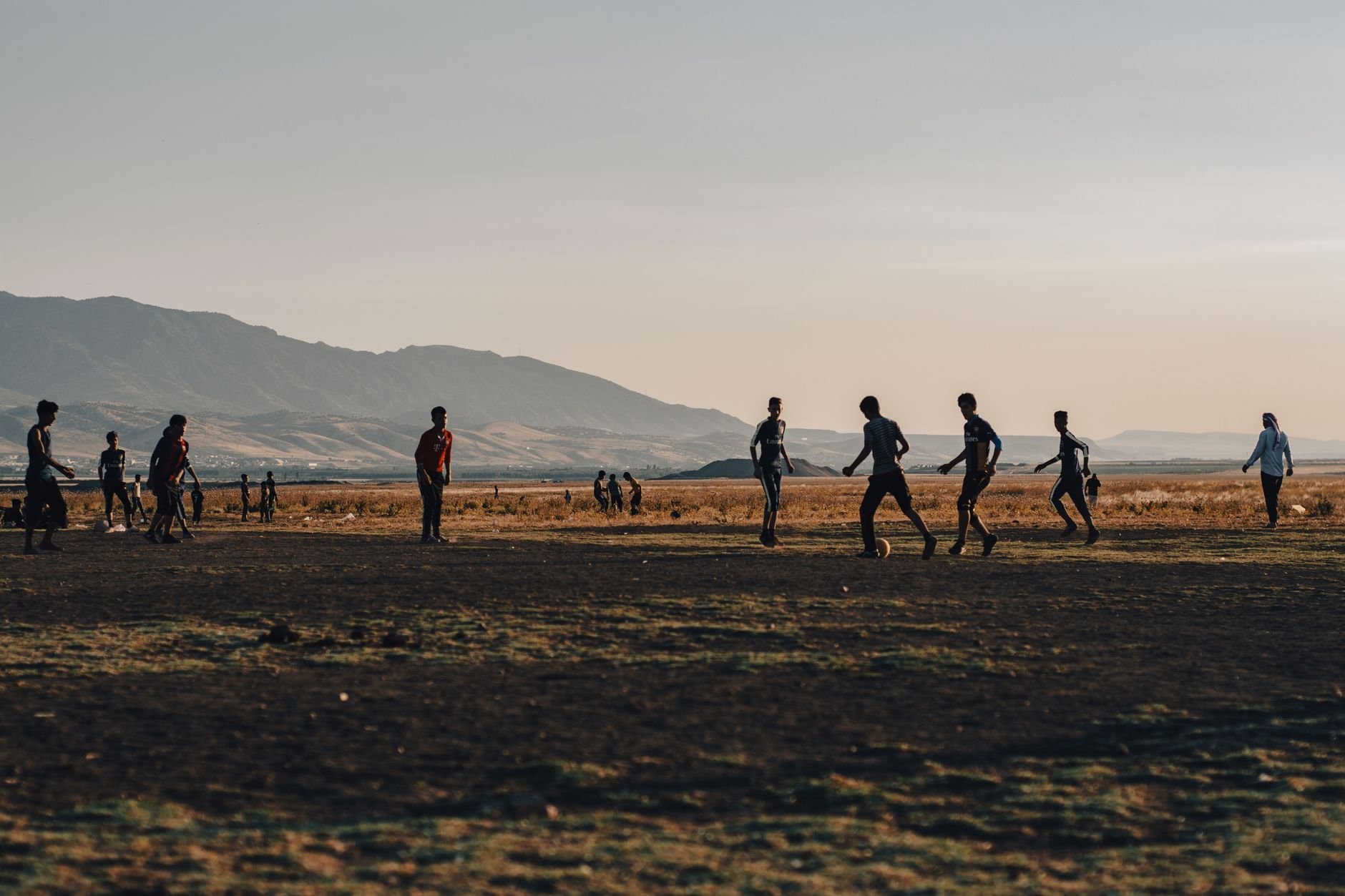 A group of people play soccer at sunset in a flat desert landscape, mountains in the background.