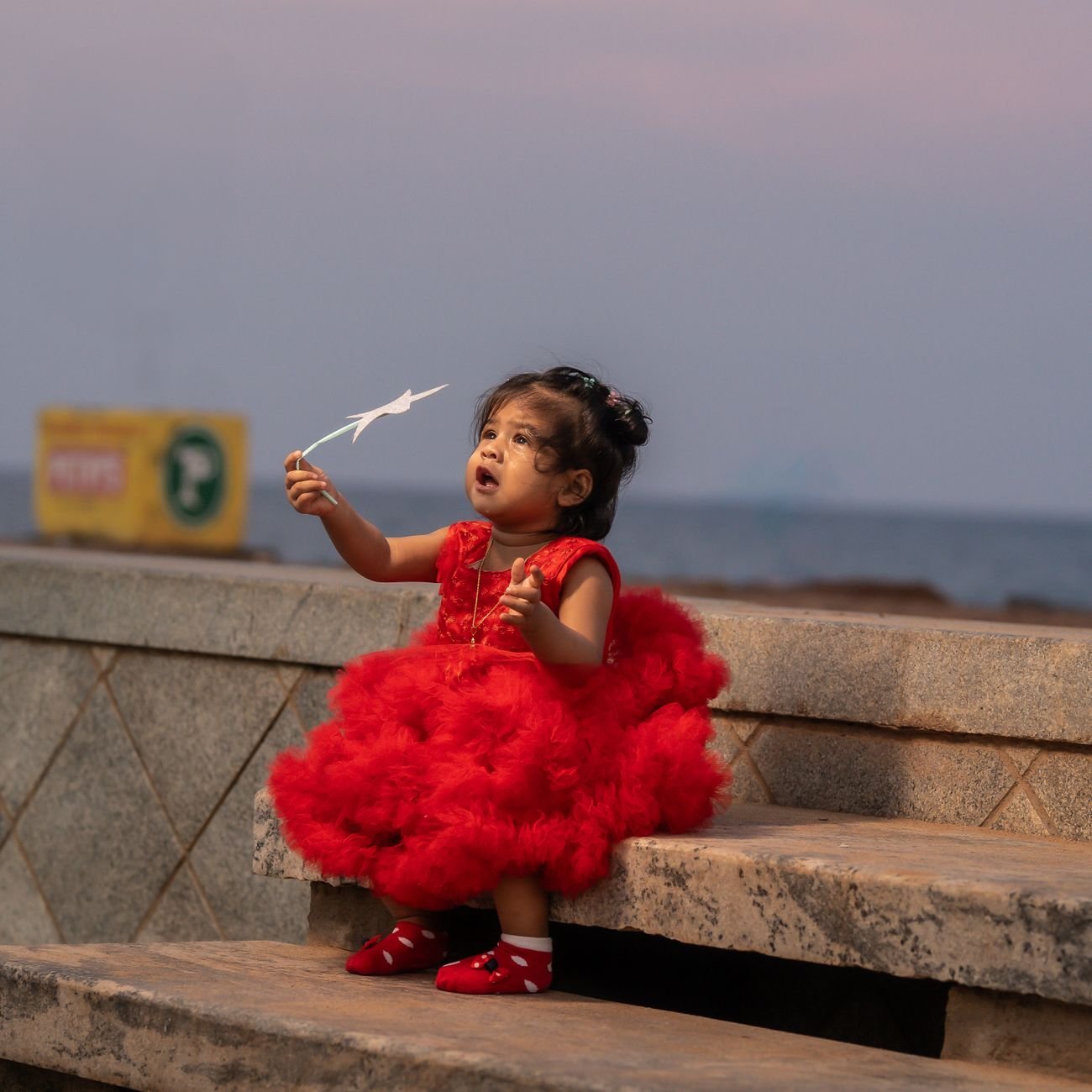 A cute child in a vibrant red dress sits on a bench by the sea at twilight, immersed in play.
