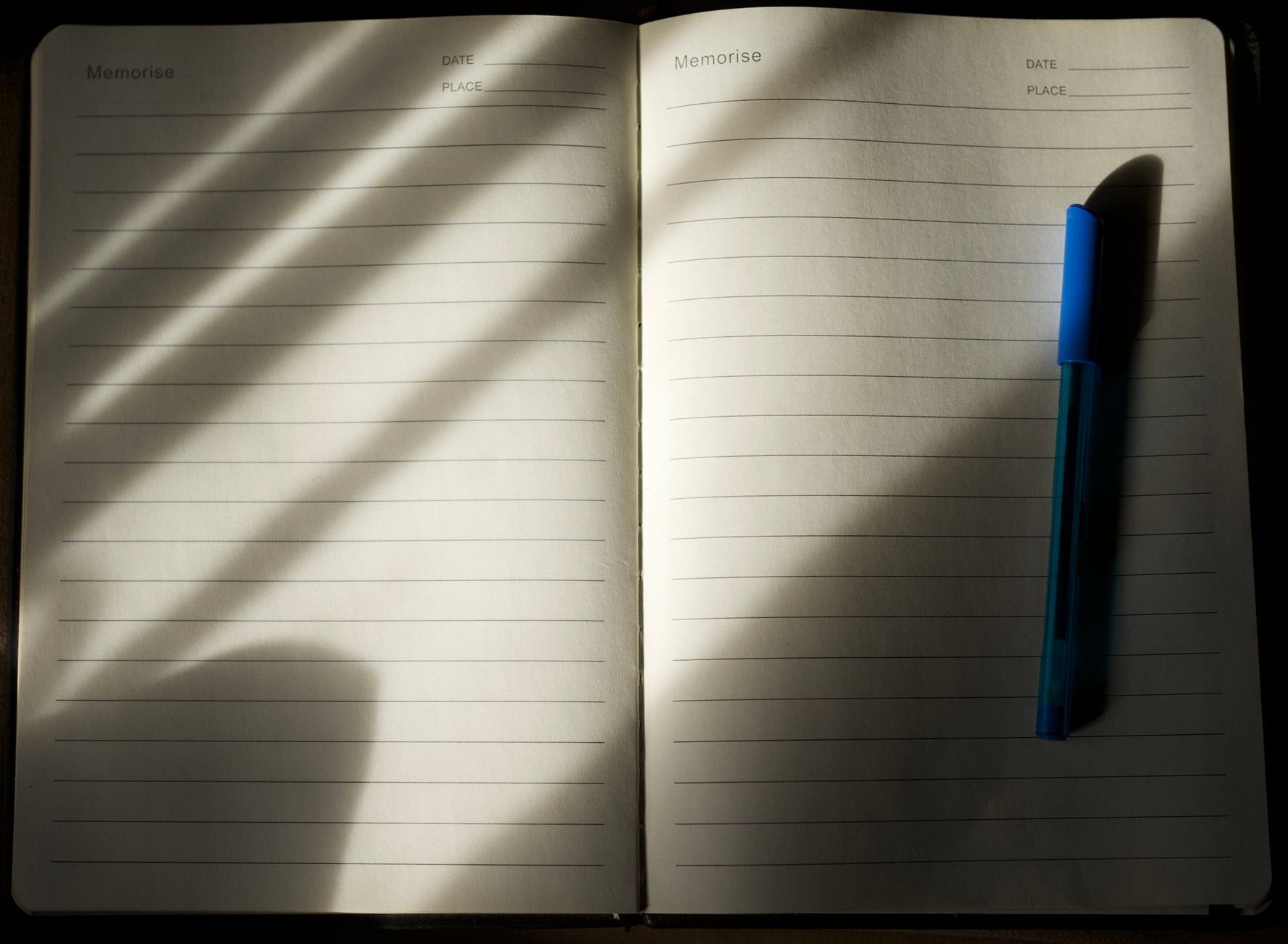 An open lined notebook with a blue pen under soft natural light casting gentle shadows.