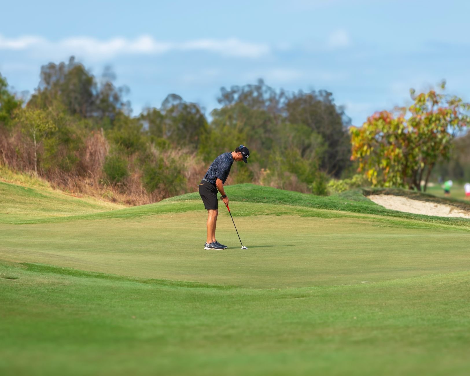 A man concentrating while putting on a lush green golf course surrounded by trees.
