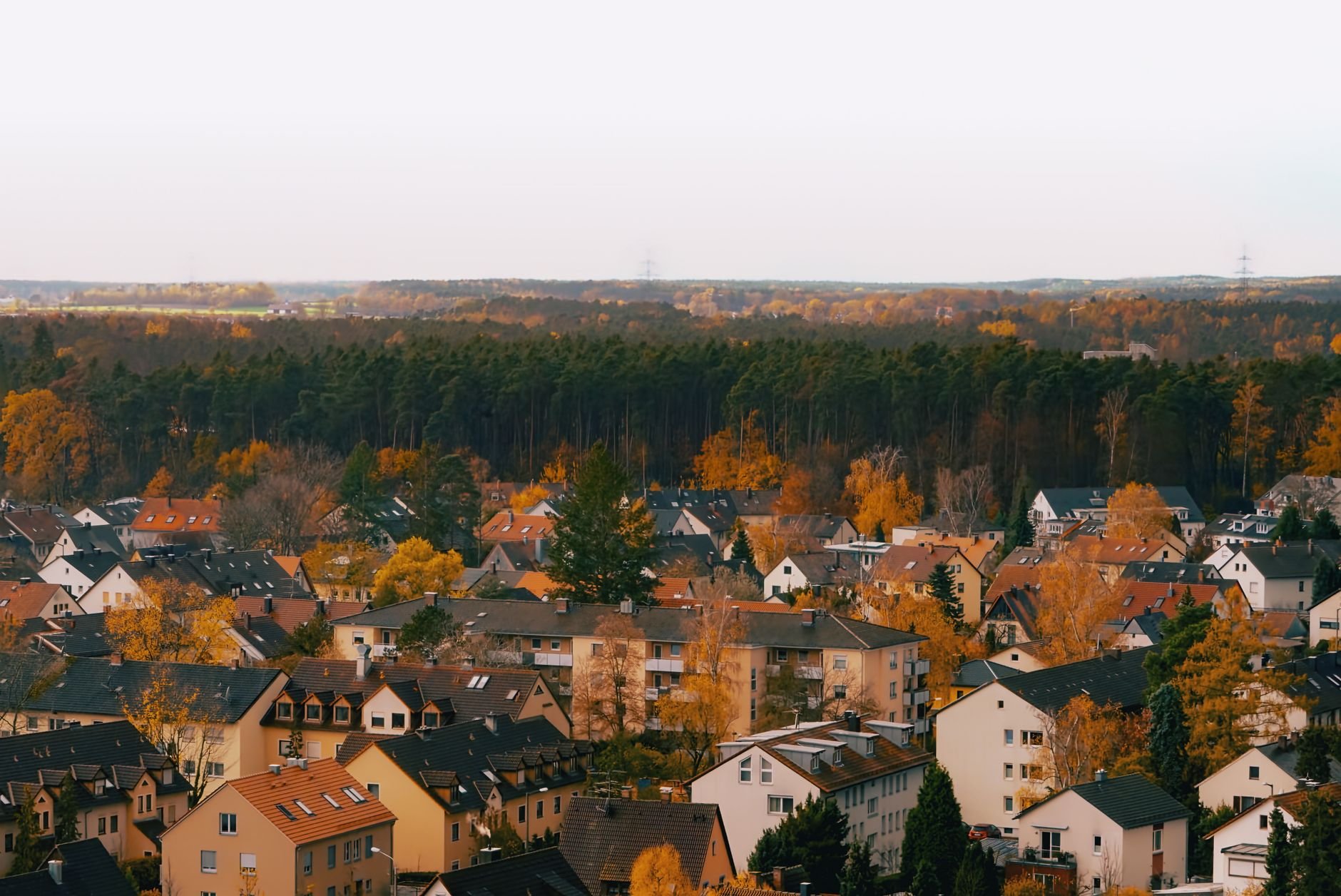 Aerial shot of a town surrounded by autumn trees, showcasing rooftops and a forest in the background.