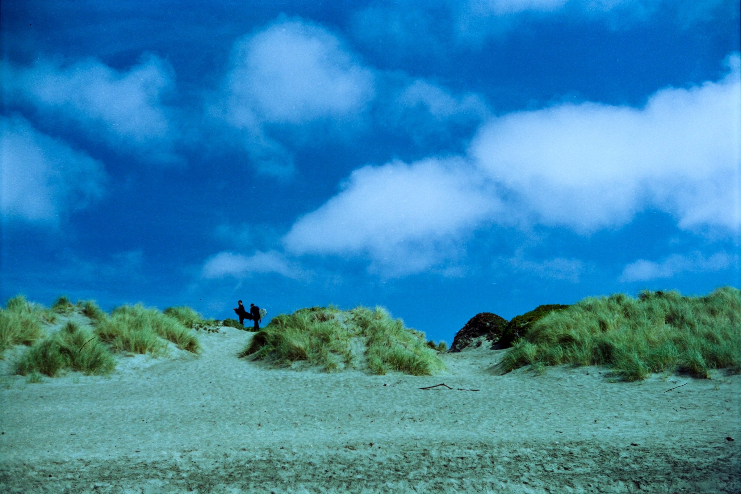 Two figures silhouetted on sand dunes beneath a dramatic blue sky, film photograph