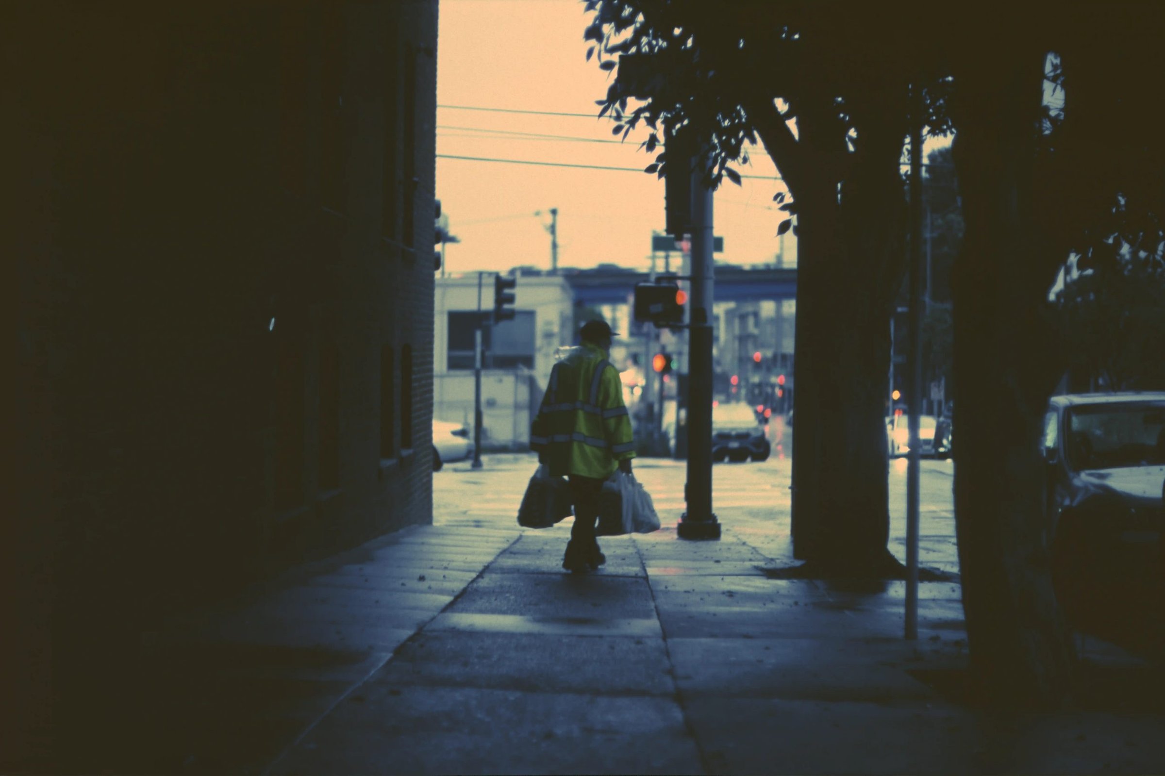 Worker in hi-vis vest carrying bags through a dark urban alley at dusk, film photograph