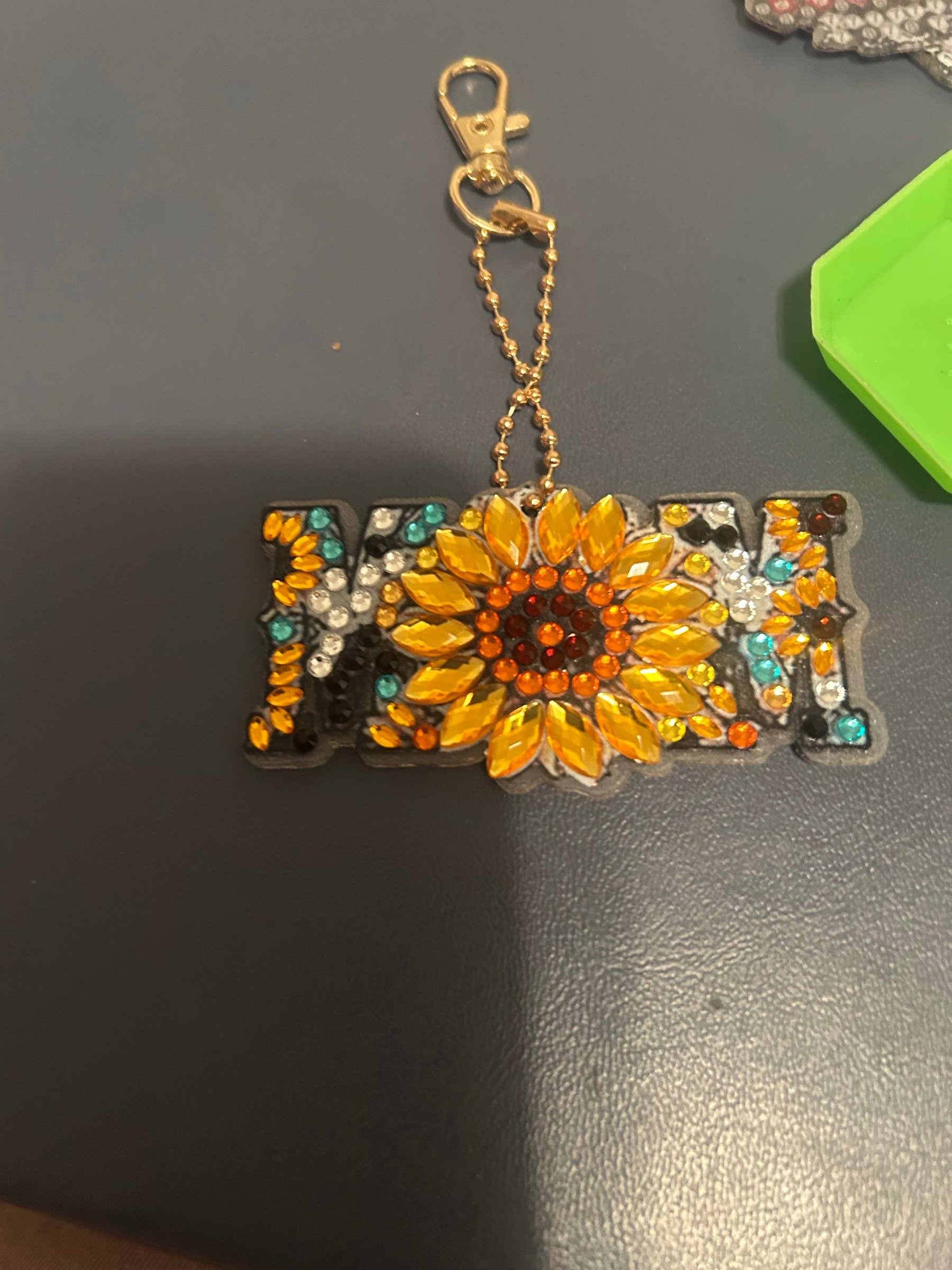 Finished diamond dot art keychain spelling MOM with a sunflower centerpiece in gold, orange, teal and white gems
